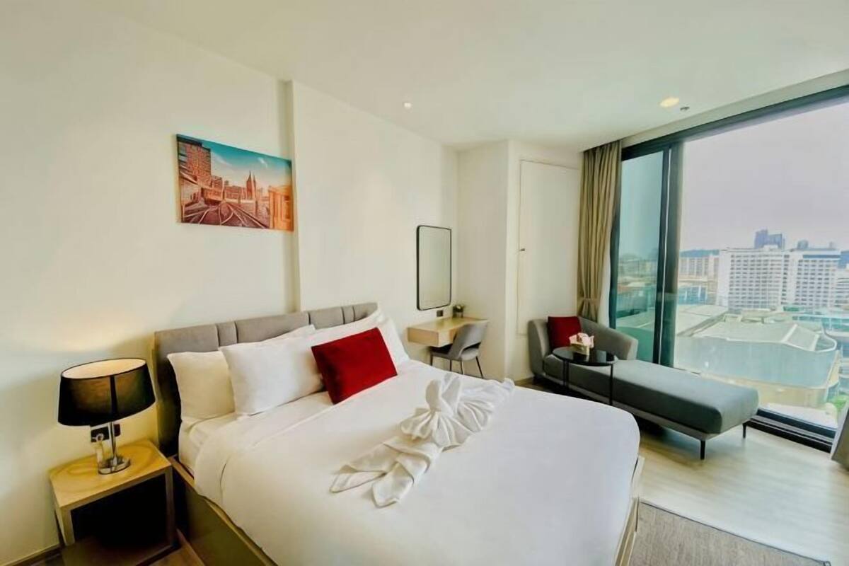 A hotel room with a large bed, a desk, a chair, a sofa, a window with a city view, and a painting on the wall.