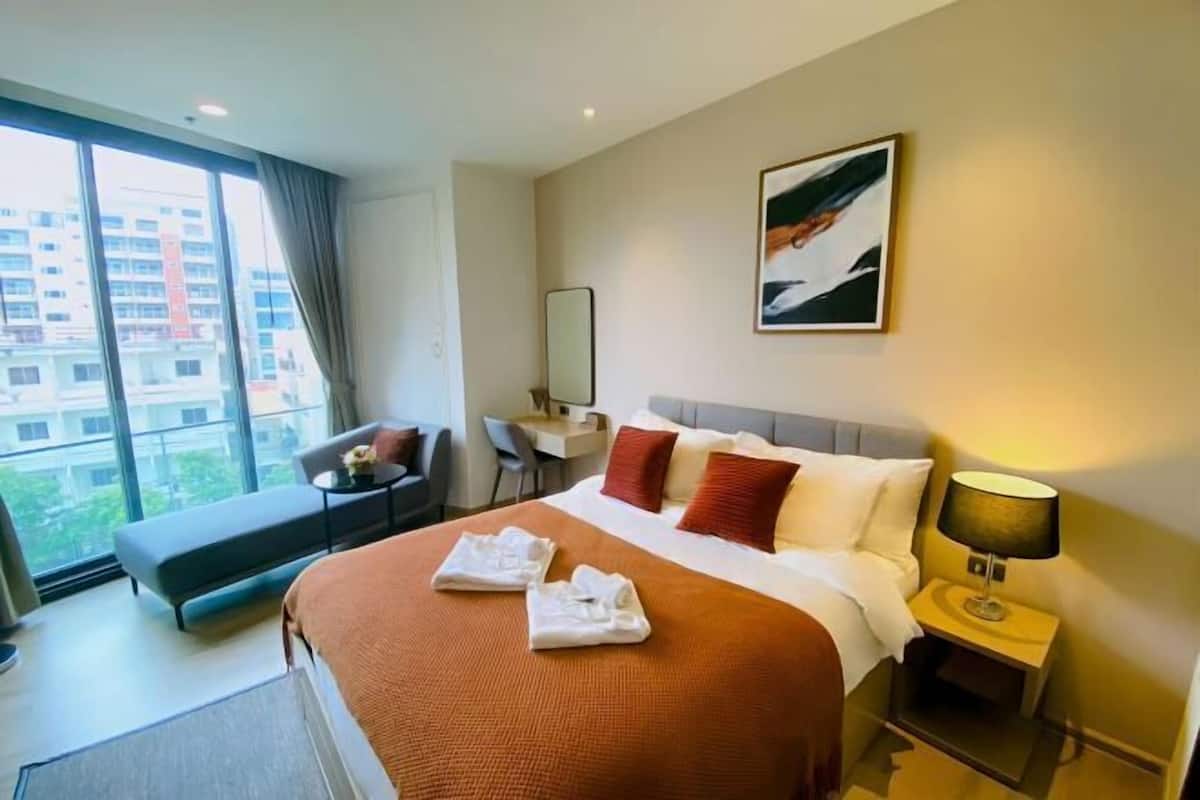 A hotel room with a large bed, a desk, a chair, and a view of buildings through the window.