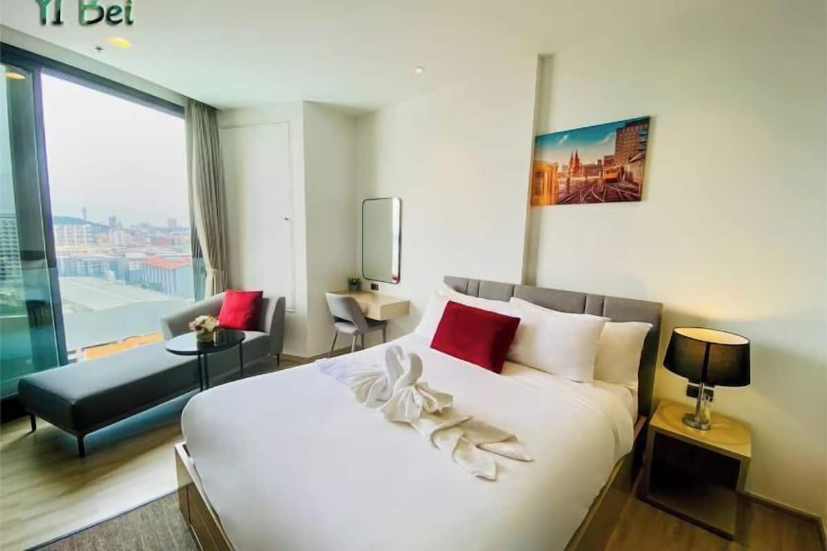 A hotel room with a large bed, a desk, a chair, a bench, a nightstand with a lamp, and a cityscape view.