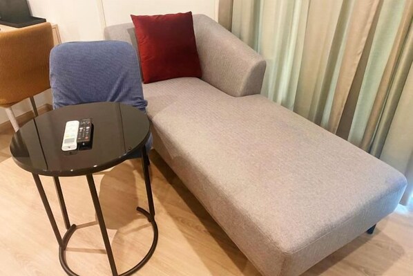 Un seul canapé avec un coussin rouge, une petite table ronde avec une télécommande, et un rideau à rayures en arrière-plan.