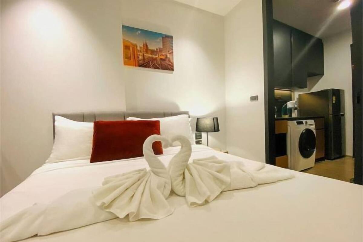 A hotel room with a bed, a red pillow, a lamp, a washing machine, and a painting on the wall.