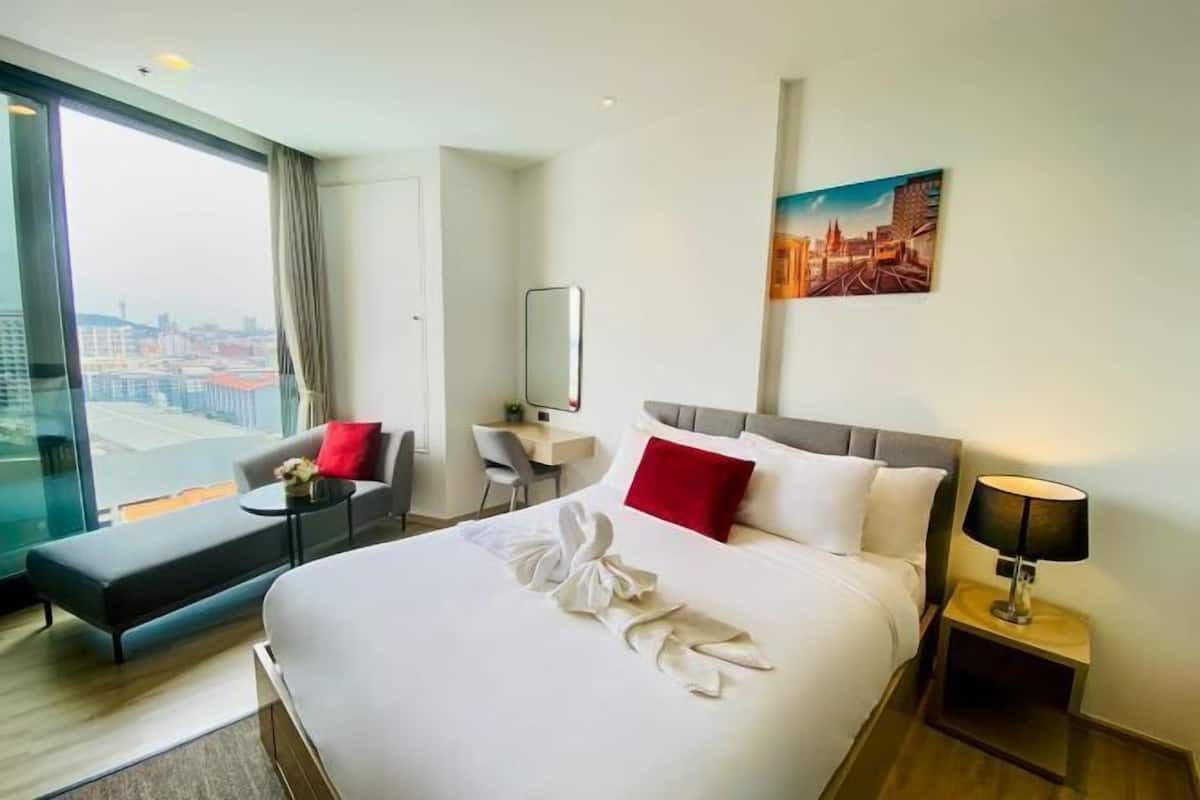 A hotel room with a large bed, a desk, a chair, a bench, a nightstand, and a city view.
