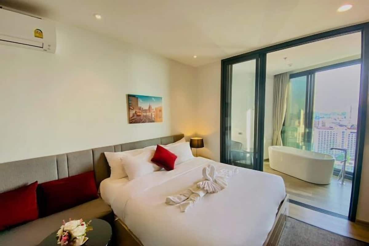 A hotel room with a bed, a sofa, a small table with flowers, a painting on the wall, and a view through a large window.