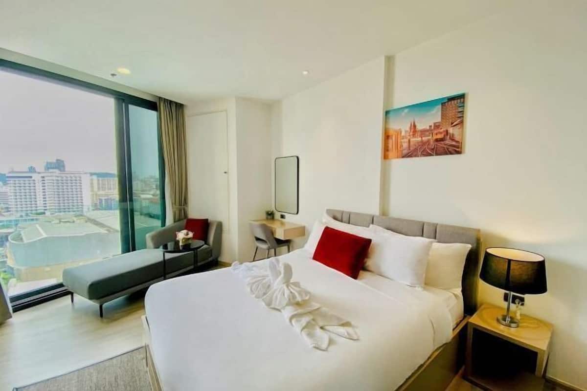 A hotel room with a large bed, a desk, a chair, a sofa, and a view of the city.