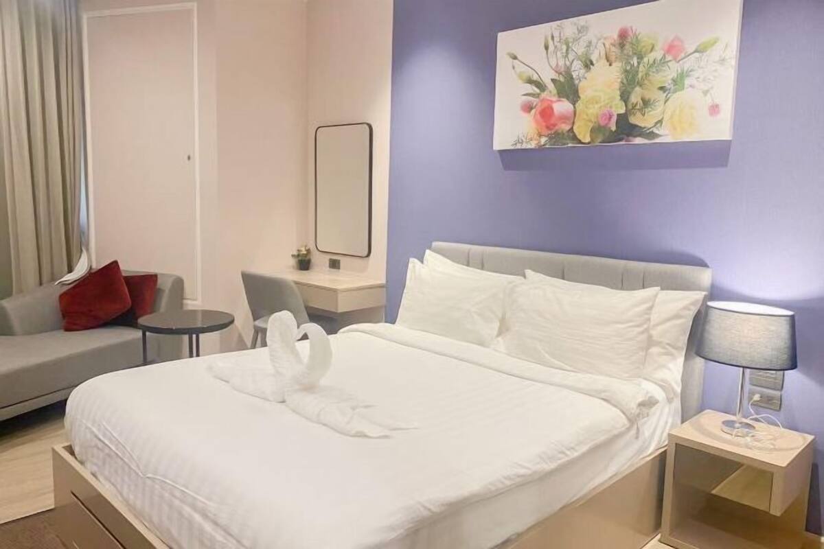 A neatly made bed with white linens, a grey headboard, a bedside table with a lamp, a small table with a chair, and a sofa with red cushions.