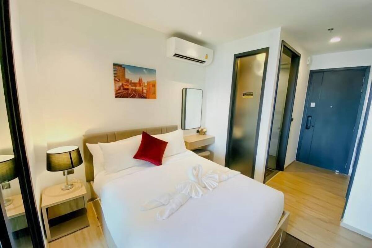 A hotel room with a bed, a nightstand, a lamp, a painting on the wall, and an air conditioning unit.