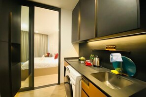 Une chambre d’hôtel de taille réduite, comprenant un lit, une petite kitchenette, un lave-linge et donnant sur la chambre voisine.
