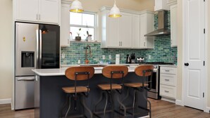 Et moderne køkken med hvide skabe, en central ø med barstole, rustfrie hvidevarer og en backsplash med grønne fliser.