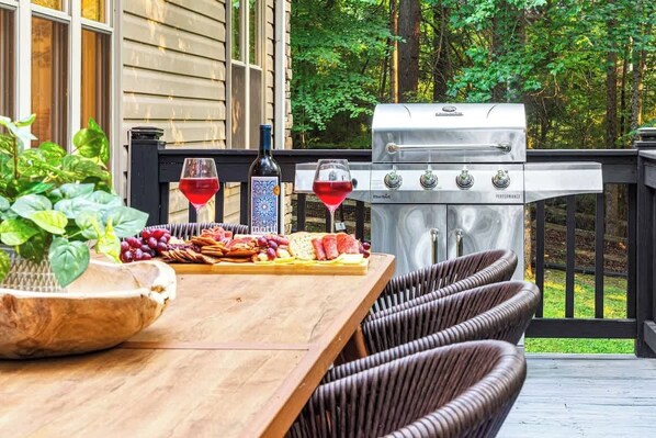 An outdoor patio with a wooden table, wicker chairs, a grill, and a glass of red wine.