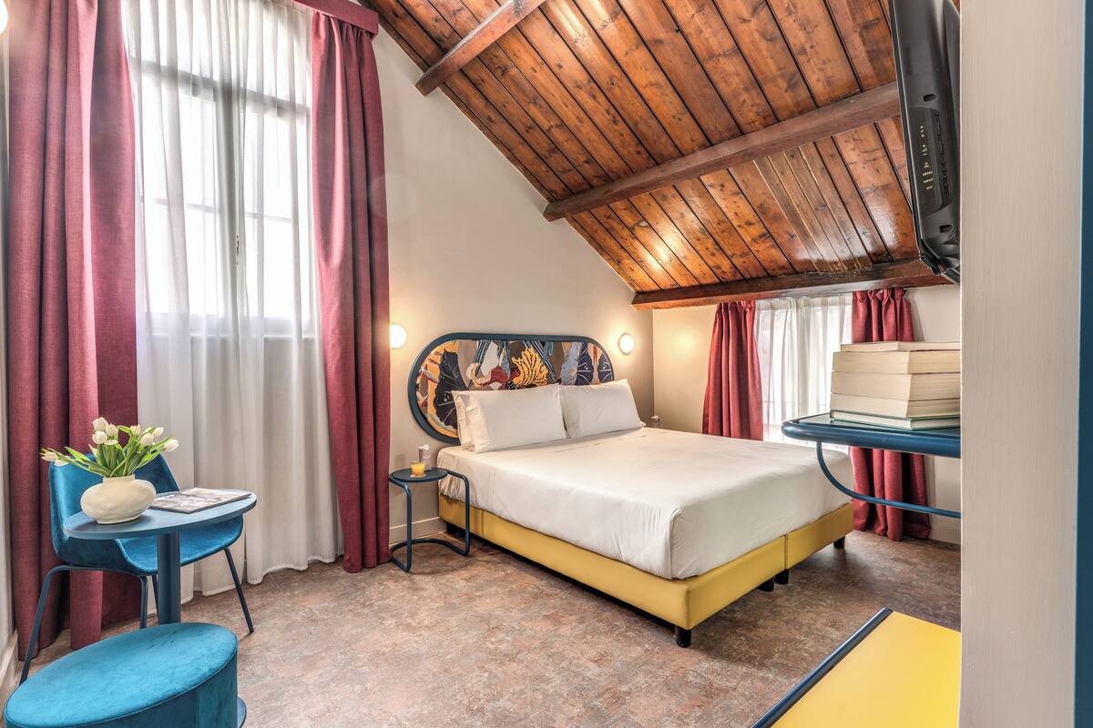 Un dormitorio con techo de madera, una cama con colchón blanco, una silla azul, una mesita con un jarrón de flores y una estantería con libros.