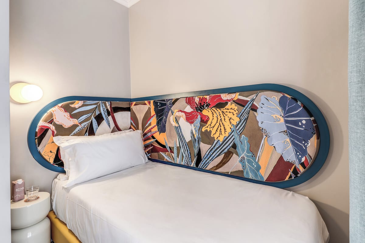 Una cama con un cabecero colorido, una mesita de noche con una caja de pañuelos y una lámpara de pared.