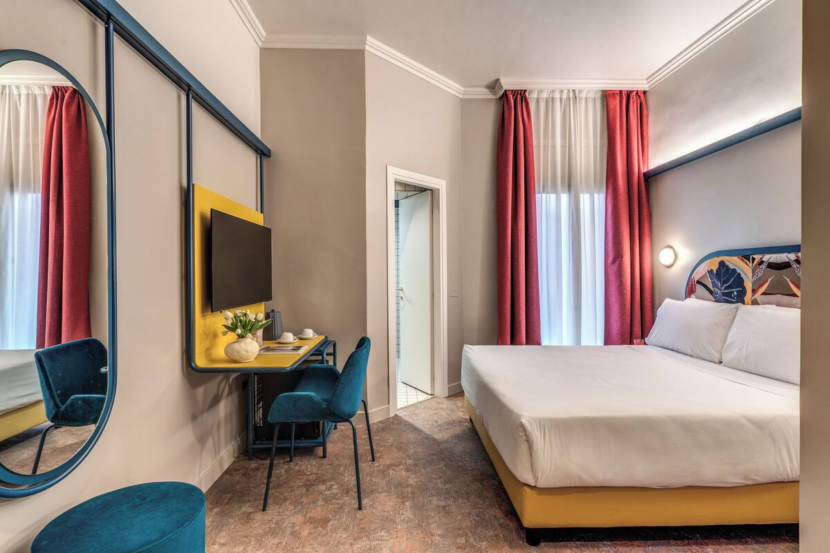 Una habitación de hotel con cama, escritorio, silla, televisor y espejo.