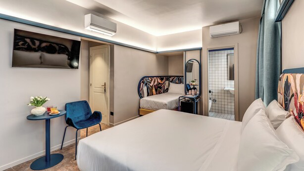 A hotel room with a large bed, a small desk with a chair, a round table with a vase and drink, and a bathroom visible through an open door. - HOTEL XX SETTEMBRE (Rome)