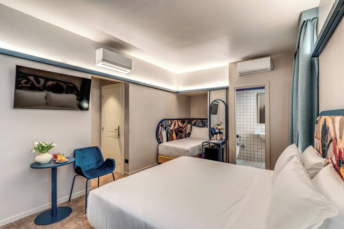 Habitación de hotel con una cama grande, un escritorio pequeño con una silla, una mesa redonda con un florero y una bebida, y un baño visible a través de una puerta abierta.
