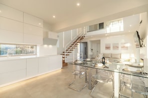 A modern kitchen with a glass dining table, clear chairs, and a staircase leading to an upper level.
