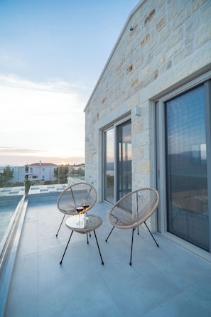 A balcony with two wicker chairs and a small table, overlooking a residential area and the sea.