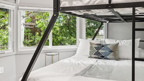 A bunk bed with white bedding and a patterned pillow, large windows with a view of greenery, and a white ceiling.