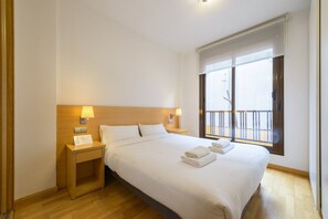 A hotel room with a large bed, wooden headboard, two bedside tables with lamps, a window with a view of a balcony, and a wooden floor. - GuestReady Essential - Relaxing stay in Barajas (Madrid)