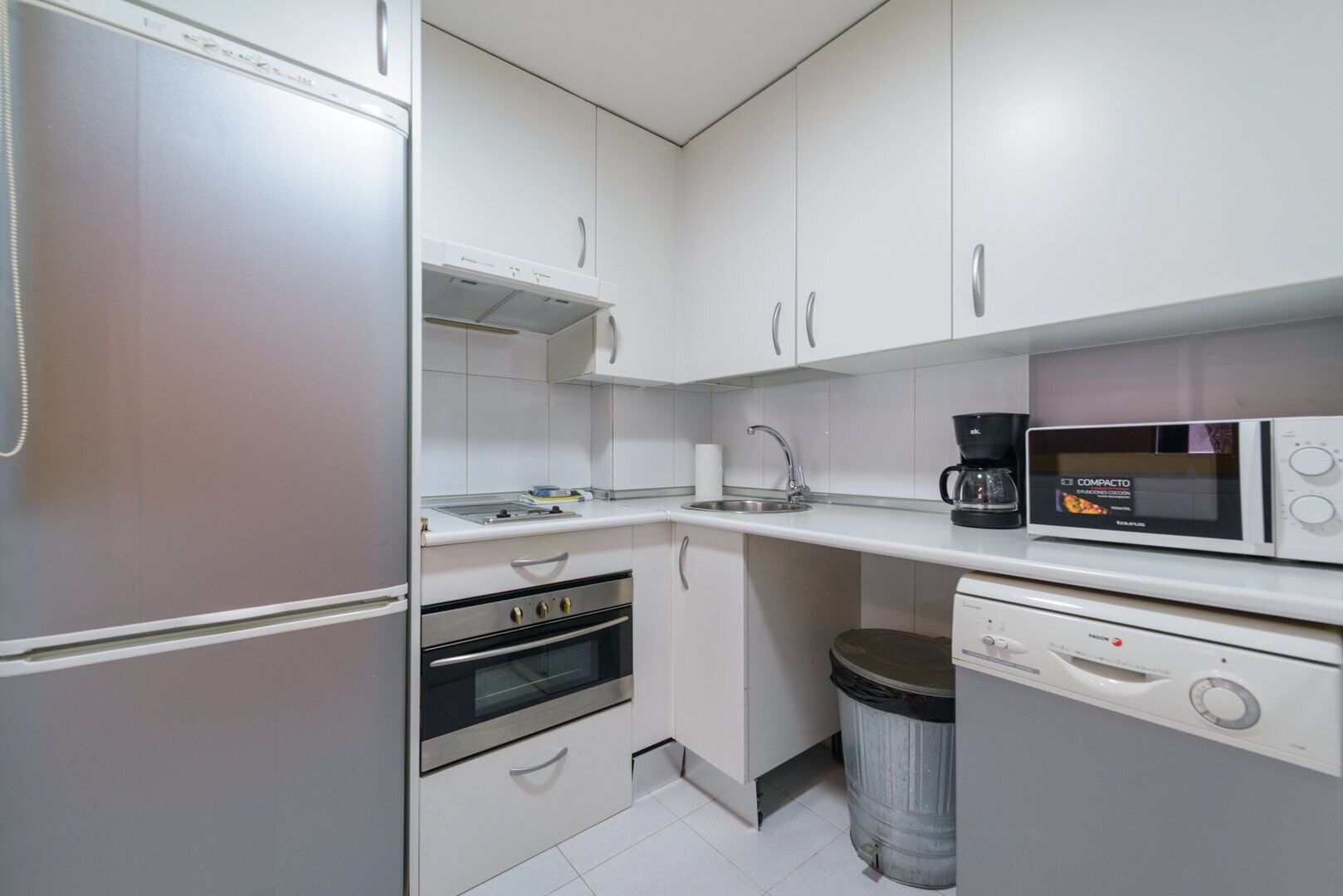 A compact kitchen with a refrigerator, oven, microwave, coffee maker, and a trash bin.