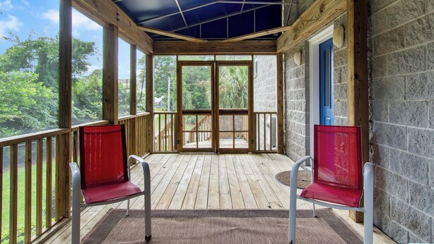 A covered porch with wooden flooring, two red chairs, and a view of greenery. - Beach Bunker | Cozy 2BR Soundside Cottage w/ Screened Porch & Fenced Yard (Emerald Isle)