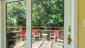 A wooden deck with a table and chairs, a grill, and a view of trees through a glass door. - Beach Bunker | Cozy 2BR Soundside Cottage w/ Screened Porch & Fenced Yard (Emerald Isle)