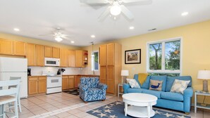 A modern kitchen with wooden cabinets, a microwave, and a stove. There is a blue patterned armchair, a blue sofa with cushions, and a white coffee table. The room has a star-patterned rug and a ceiling fan. - Beach Bunker | Cozy 2BR Soundside Cottage w/ Screened Porch & Fenced Yard (Emerald Isle)