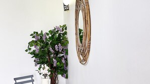 Un coin repas avec une table et des chaises, une plante en pot aux fleurs violettes, un miroir fixé au mur et un luminaire.