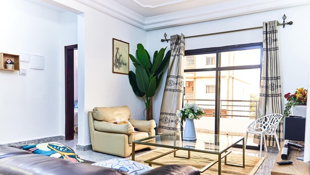 Un salon moderne avec un canapé en cuir, une table basse en verre et un fauteuil beige. On y trouve une grande plante en pot, un vase blanc avec des fleurs et une vue sur l’extérieur grâce à une grande fenêtre ornée de rideaux à motifs.