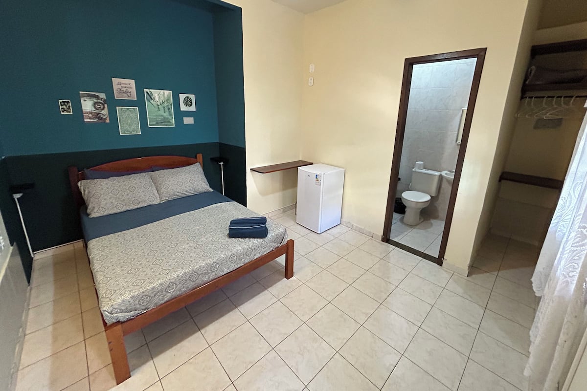 A bedroom with a bed, a small white refrigerator, a toilet, and a mirror.