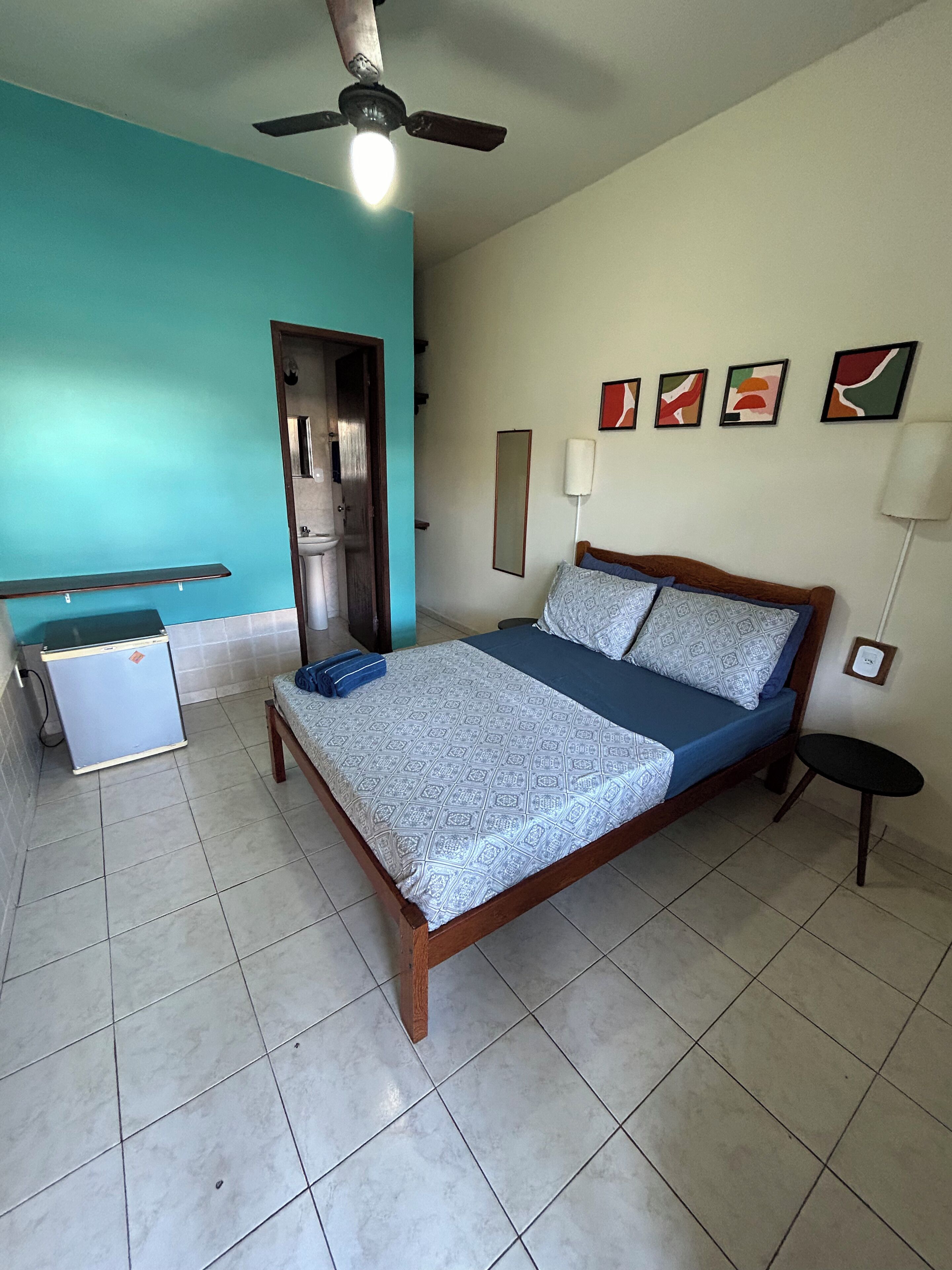 Ein Schlafzimmer mit Bett, Deckenventilator, kleinem Tisch, Stuhl, Wandlampe und einer Tür zum Badezimmer.