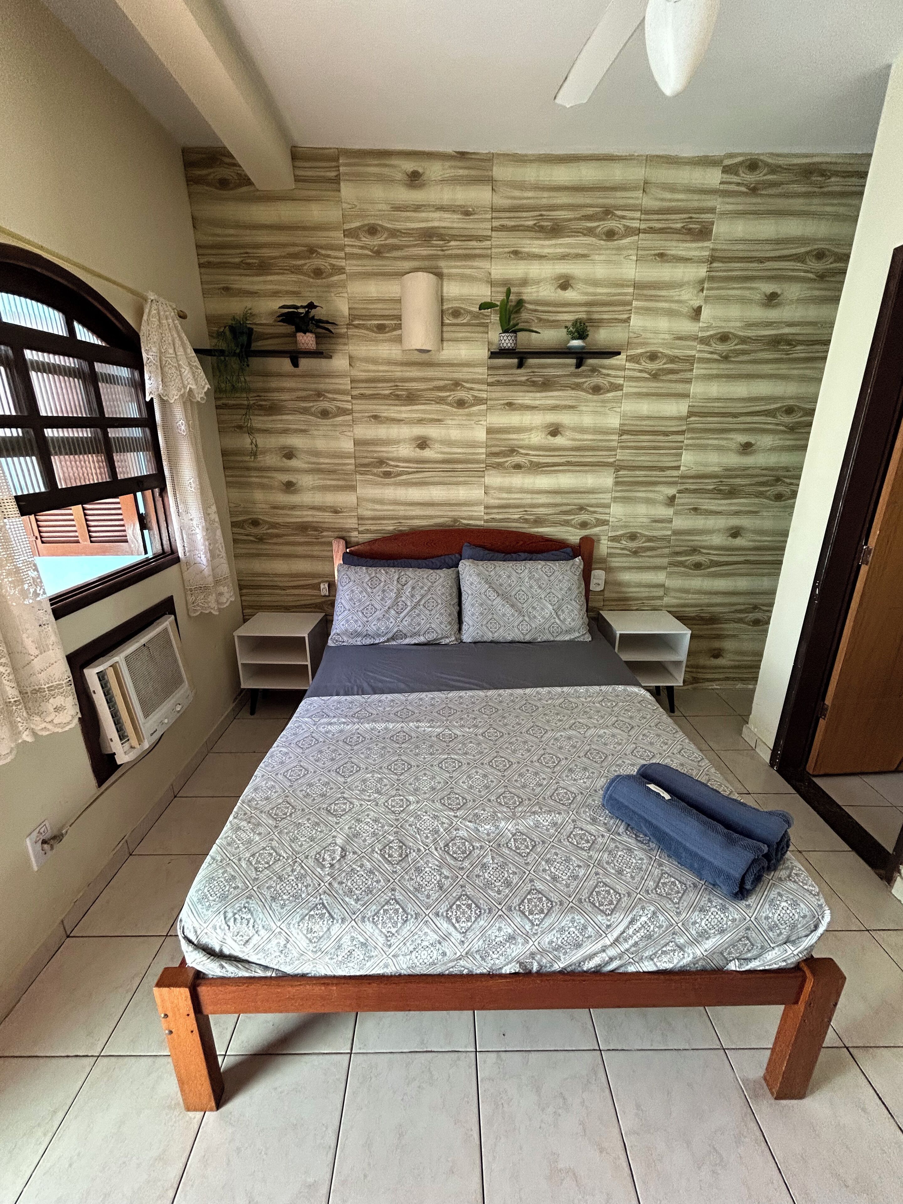 A bedroom with a wooden headboard, a bed with patterned bedding, a nightstand, a wall-mounted heater, and a window with a curtain.