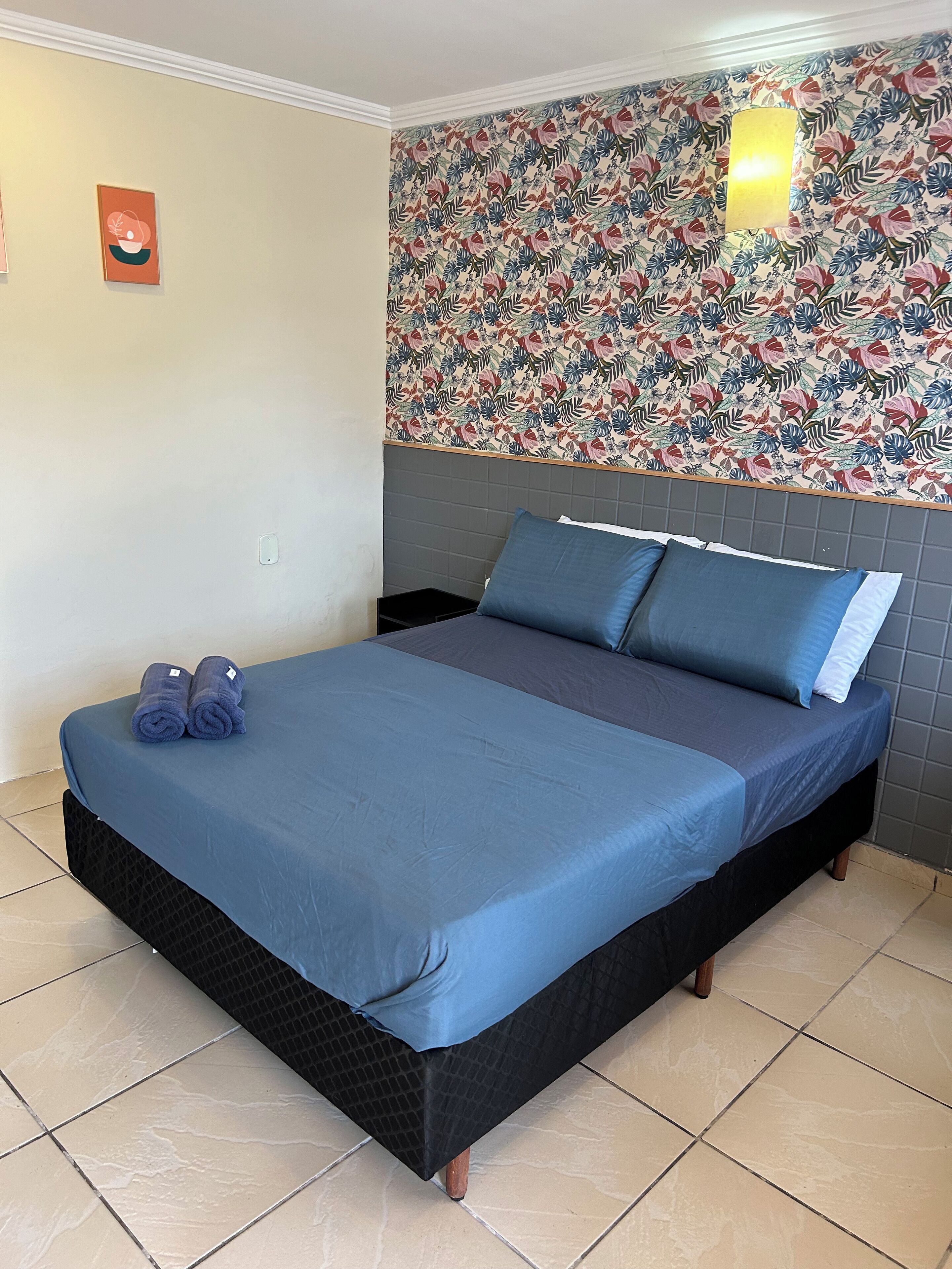 A hotel room with a bed, blue bedding, a floral wallpaper, and a small wall-mounted light.