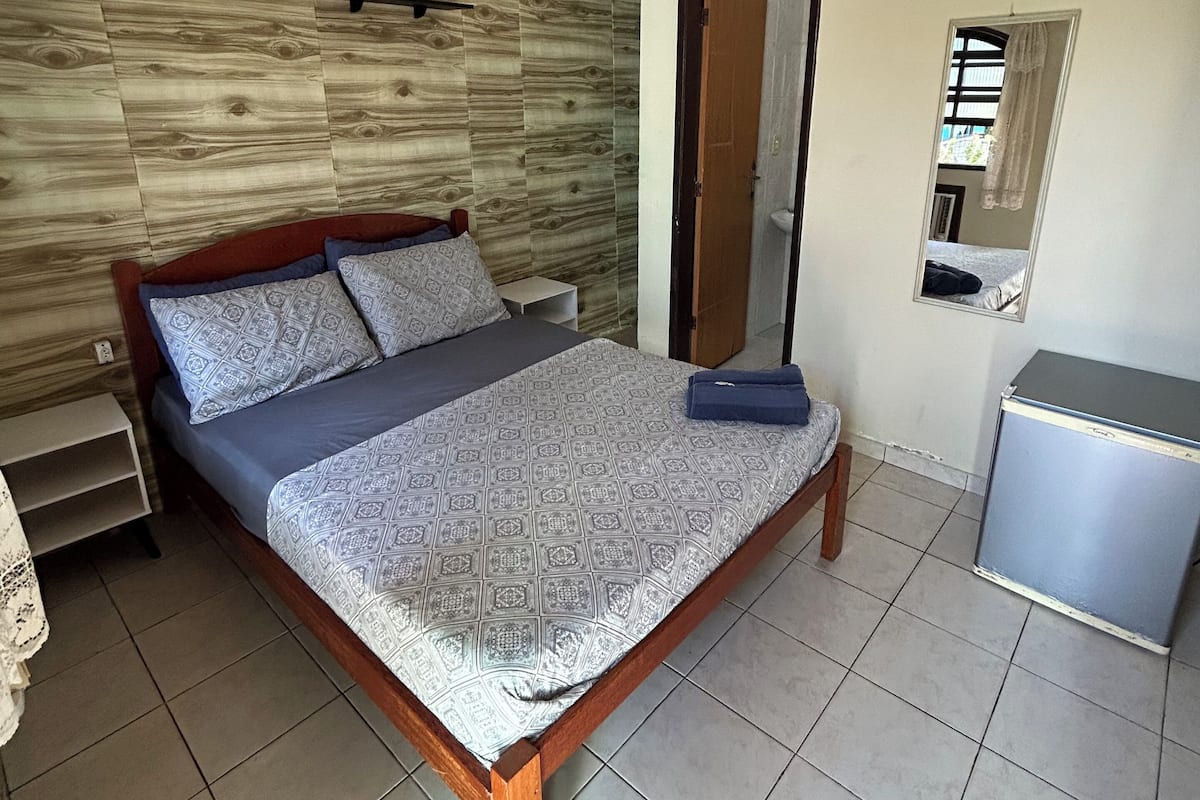 A bedroom with a bed, a ceiling fan, a mirror, a small cabinet, and a wooden wall panel.