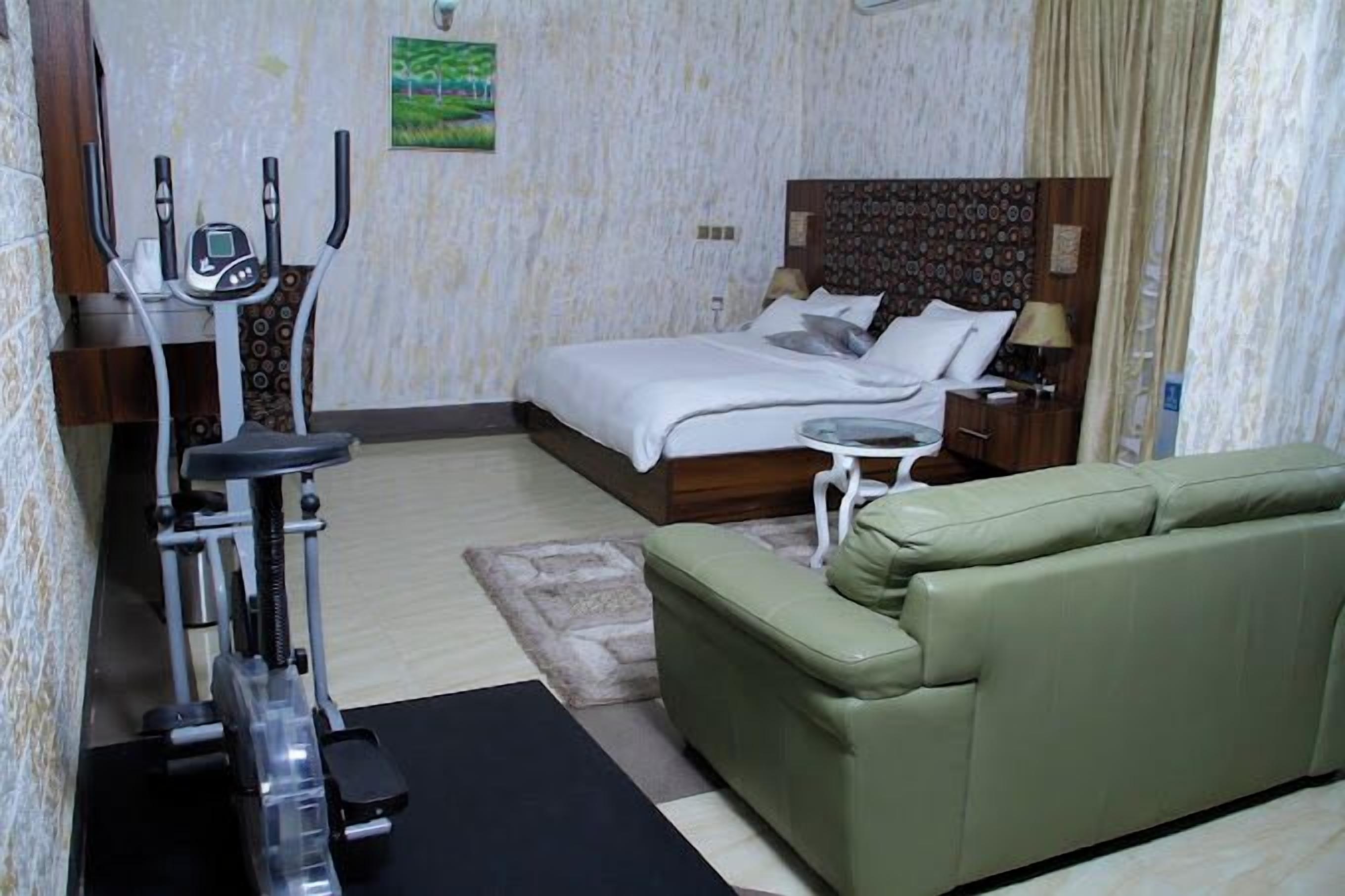 A hotel room with a bed, a green sofa, an exercise machine, a small table, and a nightstand.