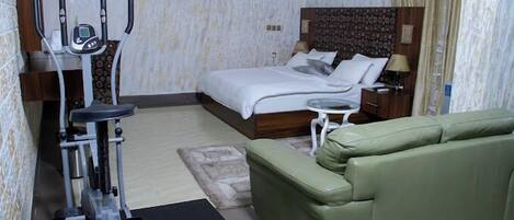 A hotel room with a bed, a green sofa, an exercise machine, a small table, and a nightstand.