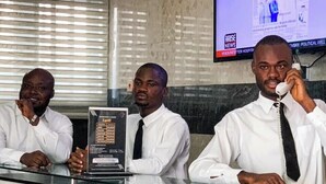 Tres hombres en un mostrador de recepción, con un monitor que muestra 'RISE NEWS' al fondo.