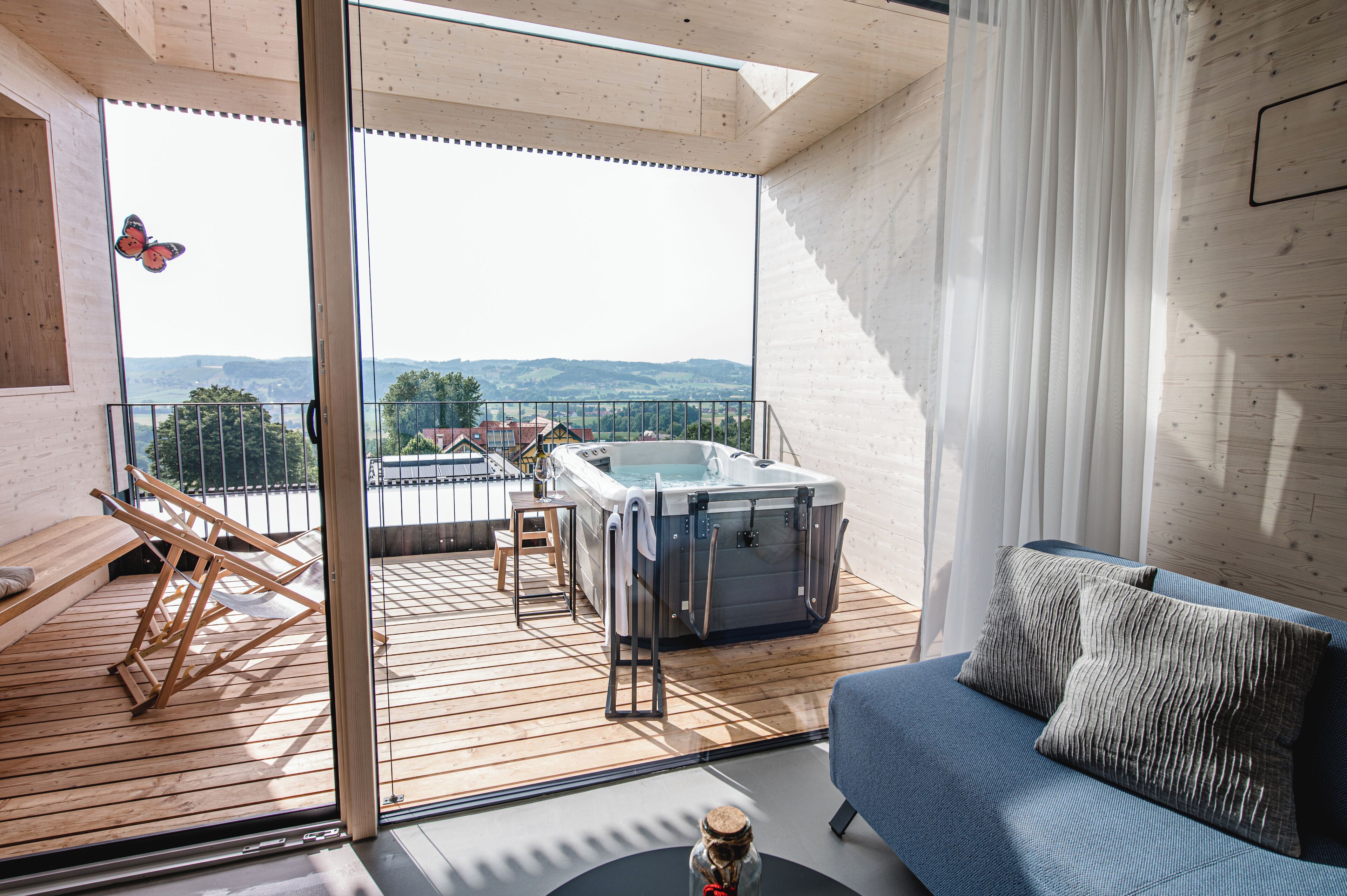 Un balcon doté d’une terrasse en bois, d’un canapé bleu, d’un jacuzzi et offrant une vue sur la campagne.
