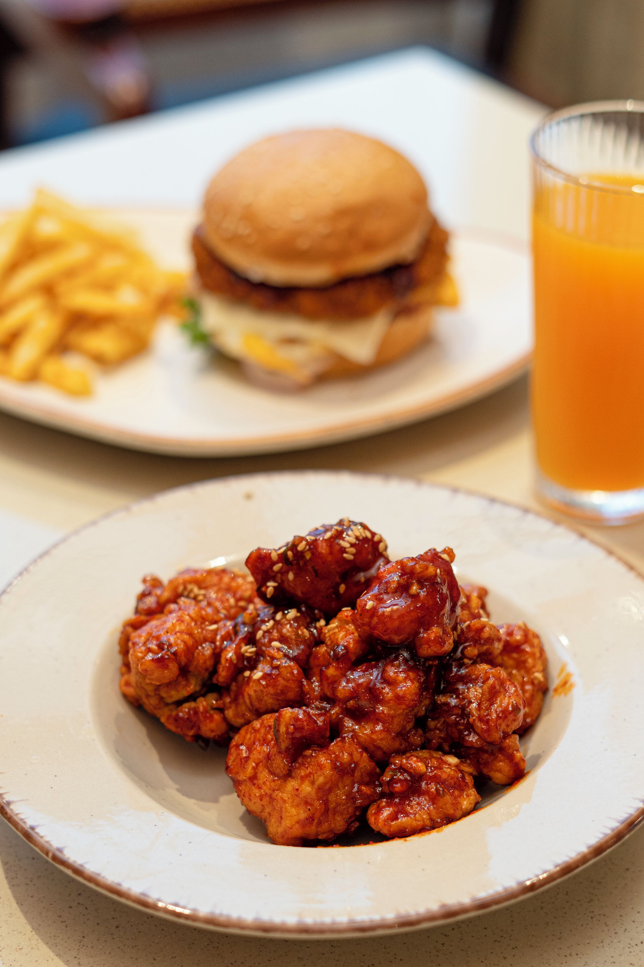 A plate of chicken wings with sesame seeds, a burger with fries, and a glass of orange juice on a table.
