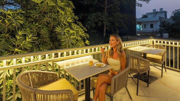 A woman dining at an outdoor table with wicker chairs and a railing, surrounded by greenery. - Sandane Homes India Expo Centre & Mart (Greater Noida)