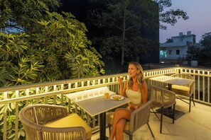 A woman dining at an outdoor table with wicker chairs and a railing, surrounded by greenery. - Sandane Homes India Expo Centre & Mart (Greater Noida)