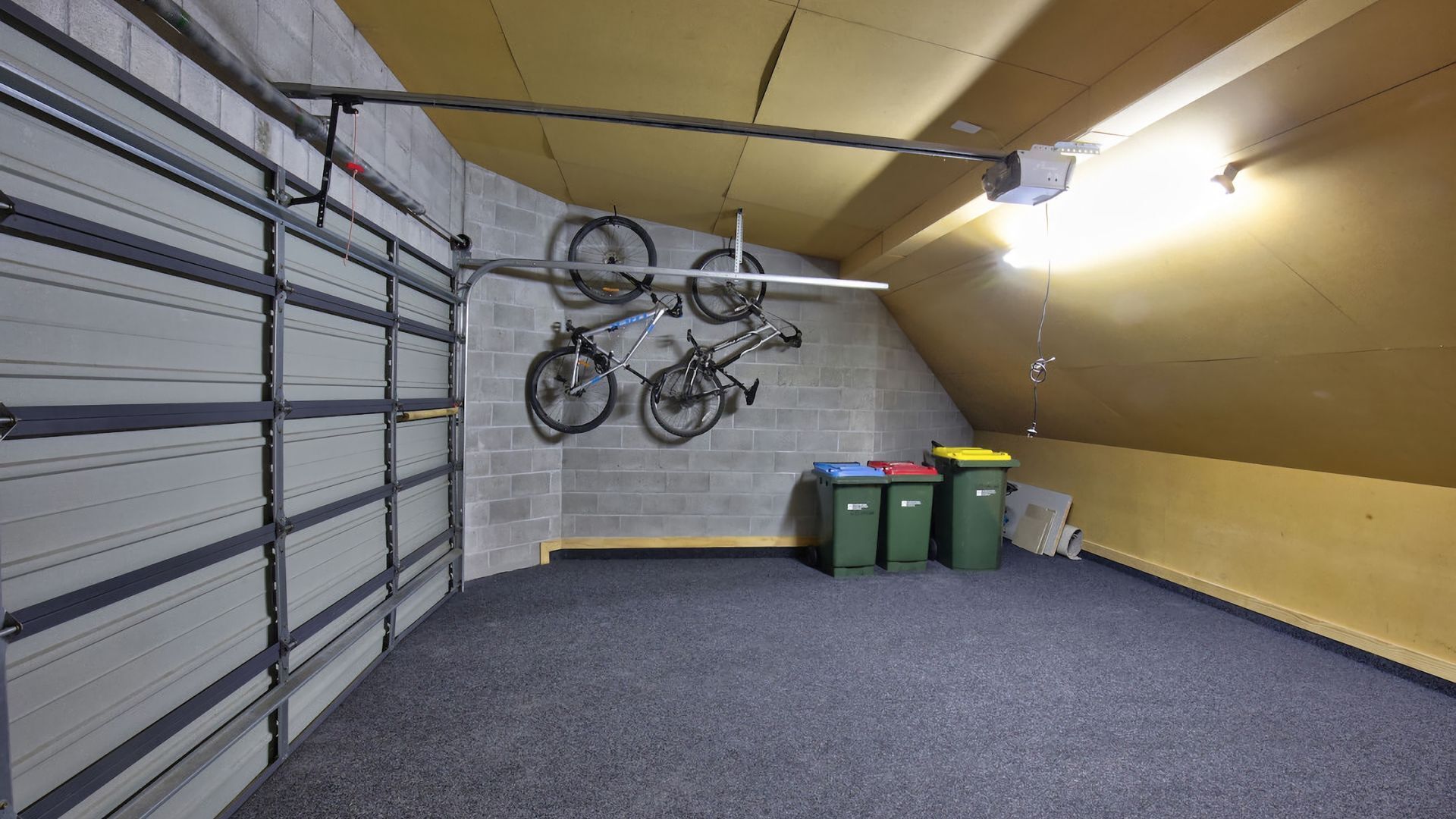Une pièce de rangement avec un plafond en pente, un porte-vélos en métal et des poubelles vertes.