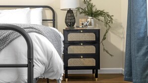 A neatly made bed with a black metal frame, a white comforter, and a patterned throw. Beside the bed is a black nightstand with three drawers and a lamp.