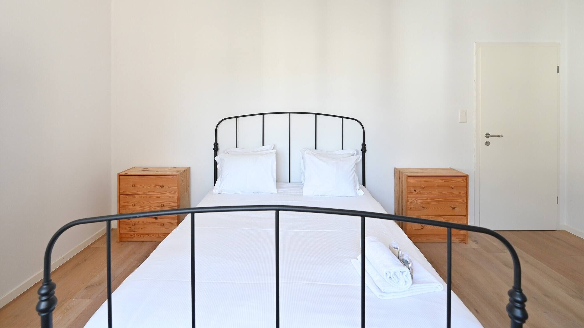 A simple, clean bedroom with a metal bed frame, white bedding, and two wooden nightstands.