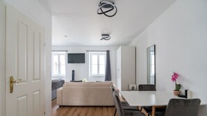 A modern living room with a sofa, a dining table with chairs, a flat-screen TV mounted on the wall, and a large window with curtains.