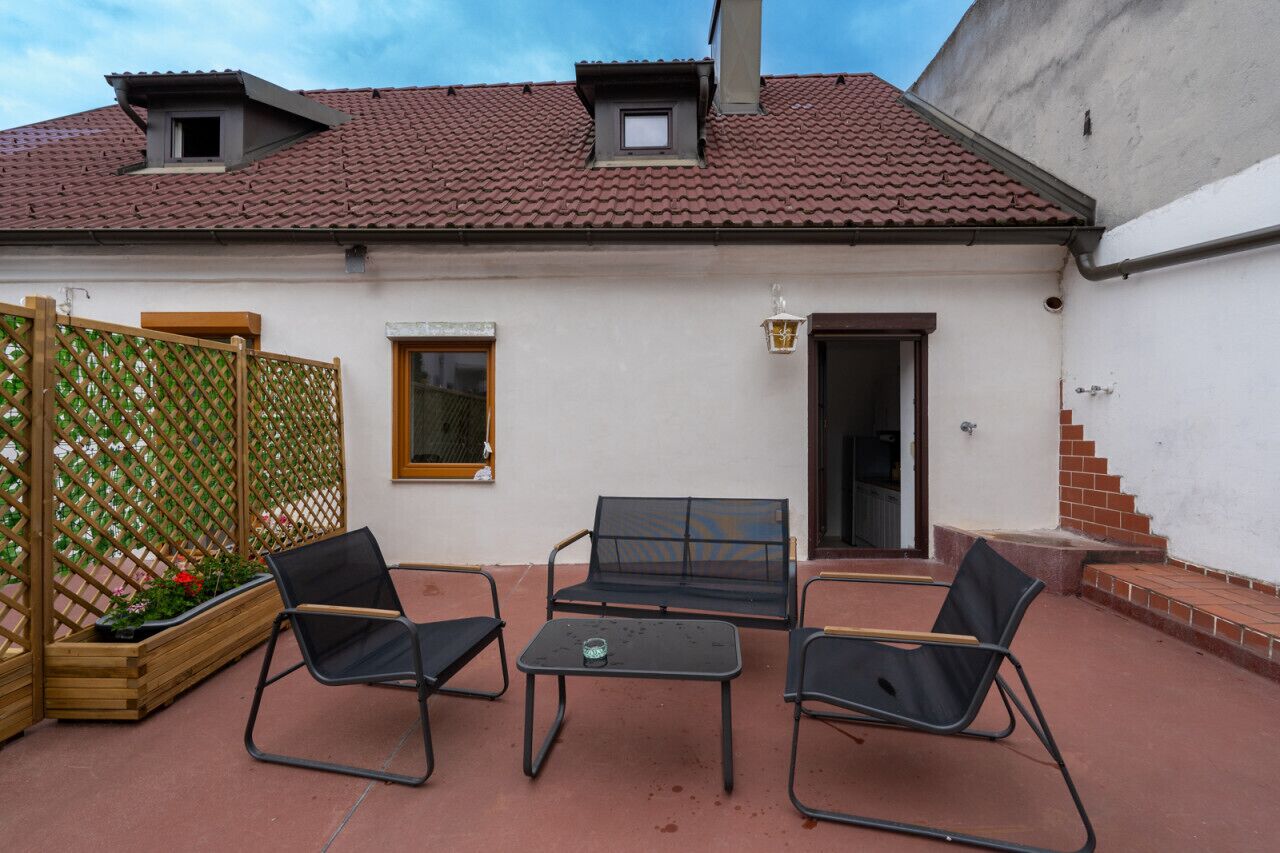 A patio with two black chairs and a table, a wooden fence with plants, and a building with a red-tiled roof and white walls.