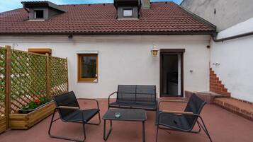 A patio with two black chairs and a table, a wooden fence with plants, and a building with a red-tiled roof and white walls.