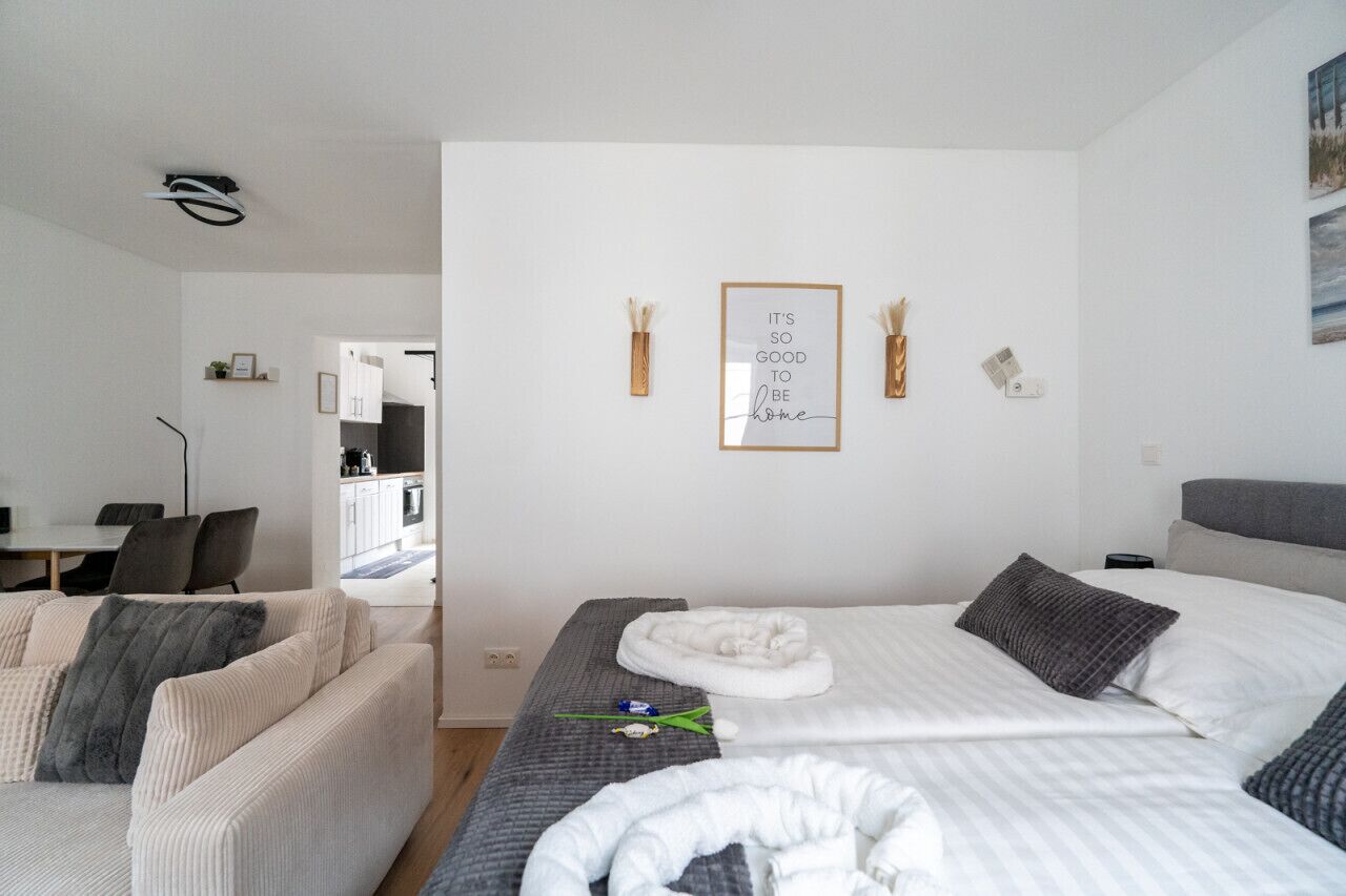 A modern hotel room with a bed, a sofa, and a desk. There are framed pictures on the wall and a small plant.