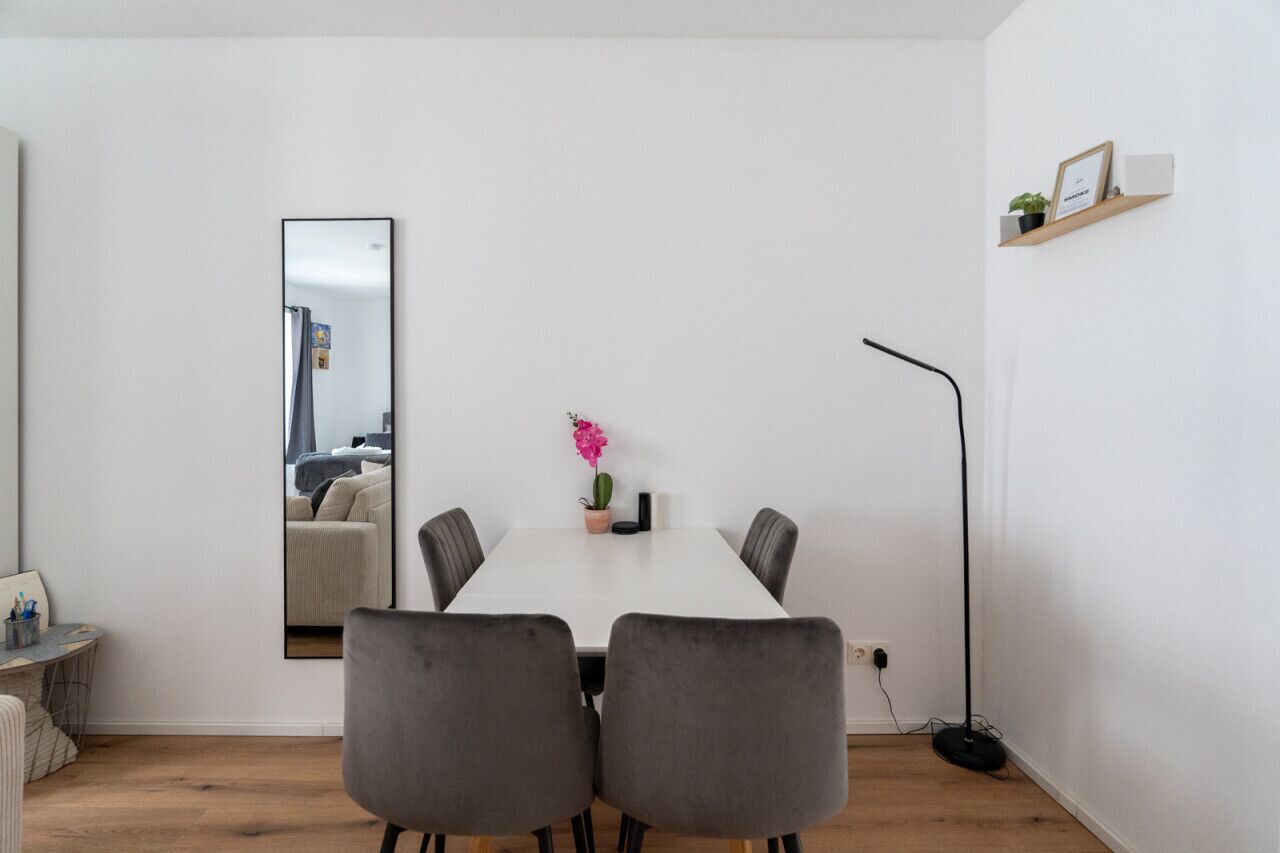 A modern, minimalist room with a white table, four grey chairs, a single potted plant, a black floor lamp, and a large mirror on the wall.