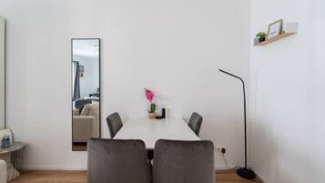 A modern, minimalist room with a white table, four grey chairs, a single potted plant, a black floor lamp, and a large mirror on the wall.