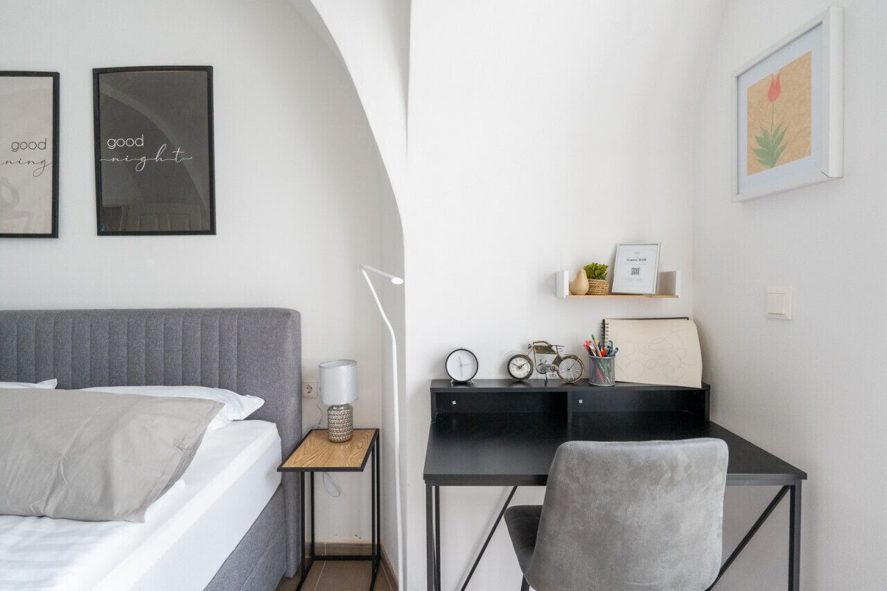 A bedroom with a bed, bedside table, lamp, and a desk with a chair, a clock, and a small plant.
